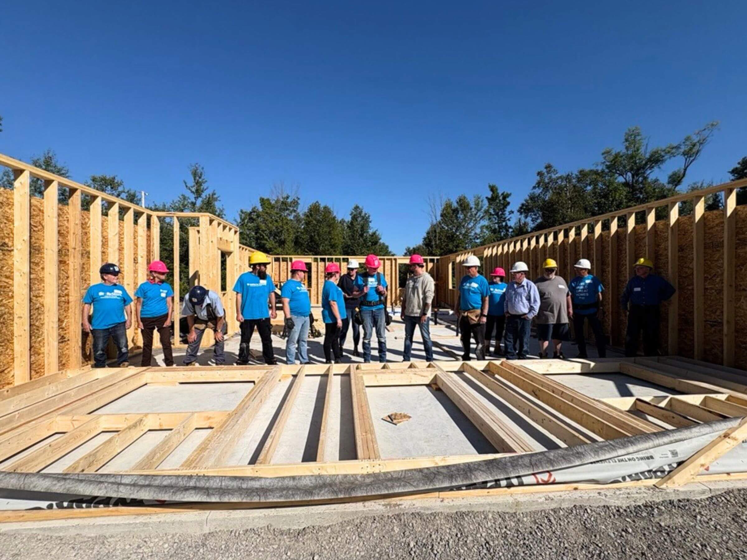 A large team of Habitat volunteers all in matching blue shirts and hardhats are standing inside an under construction home. They are all lined up in front of a framed in wall laying down, ready to be pushed up