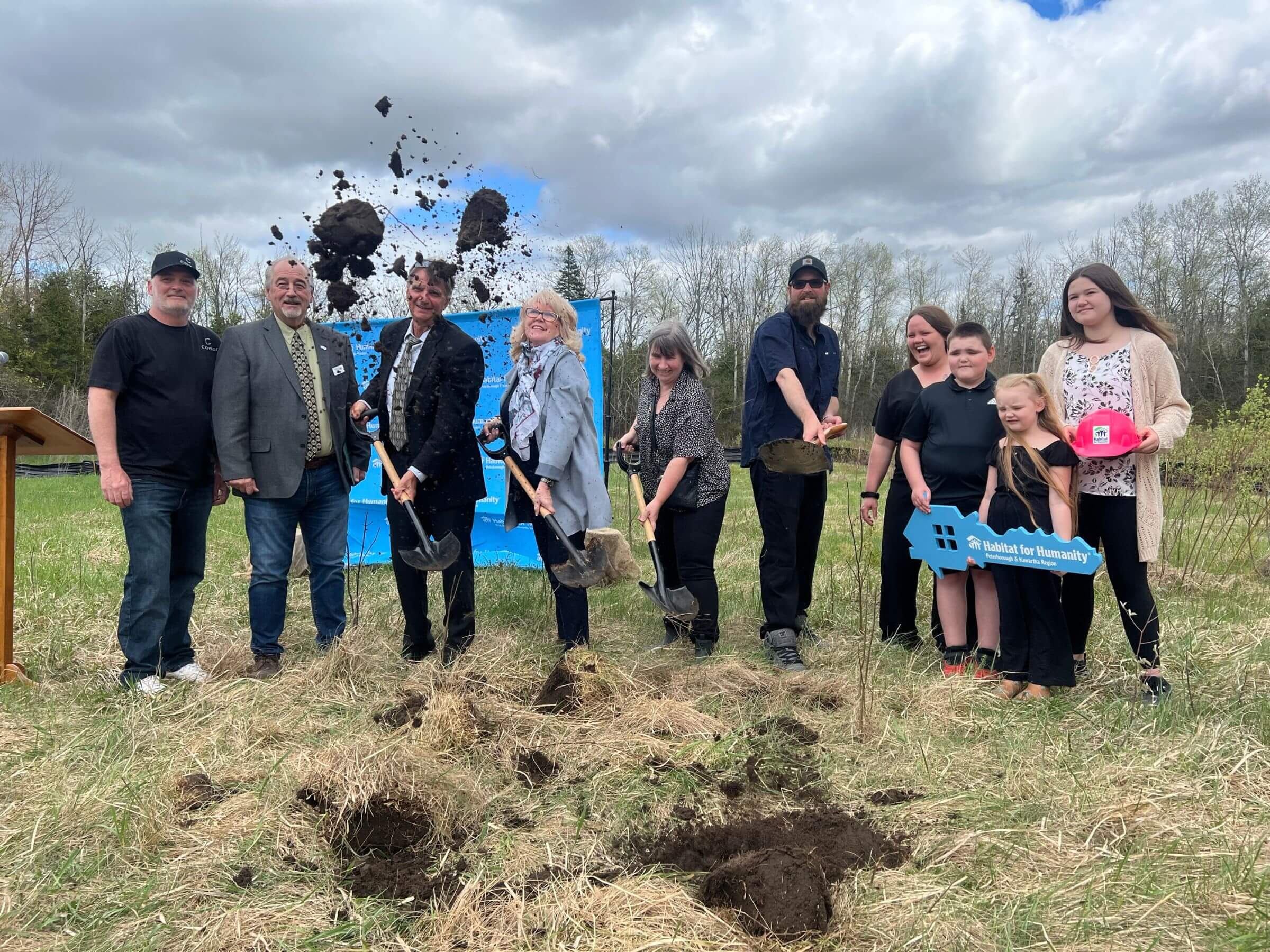 Ground is being broken outside in a field with a large blue Habitat banner in the background, a diverse group of people are holding shovels and playfully tossing dirt into the air with big smiles.