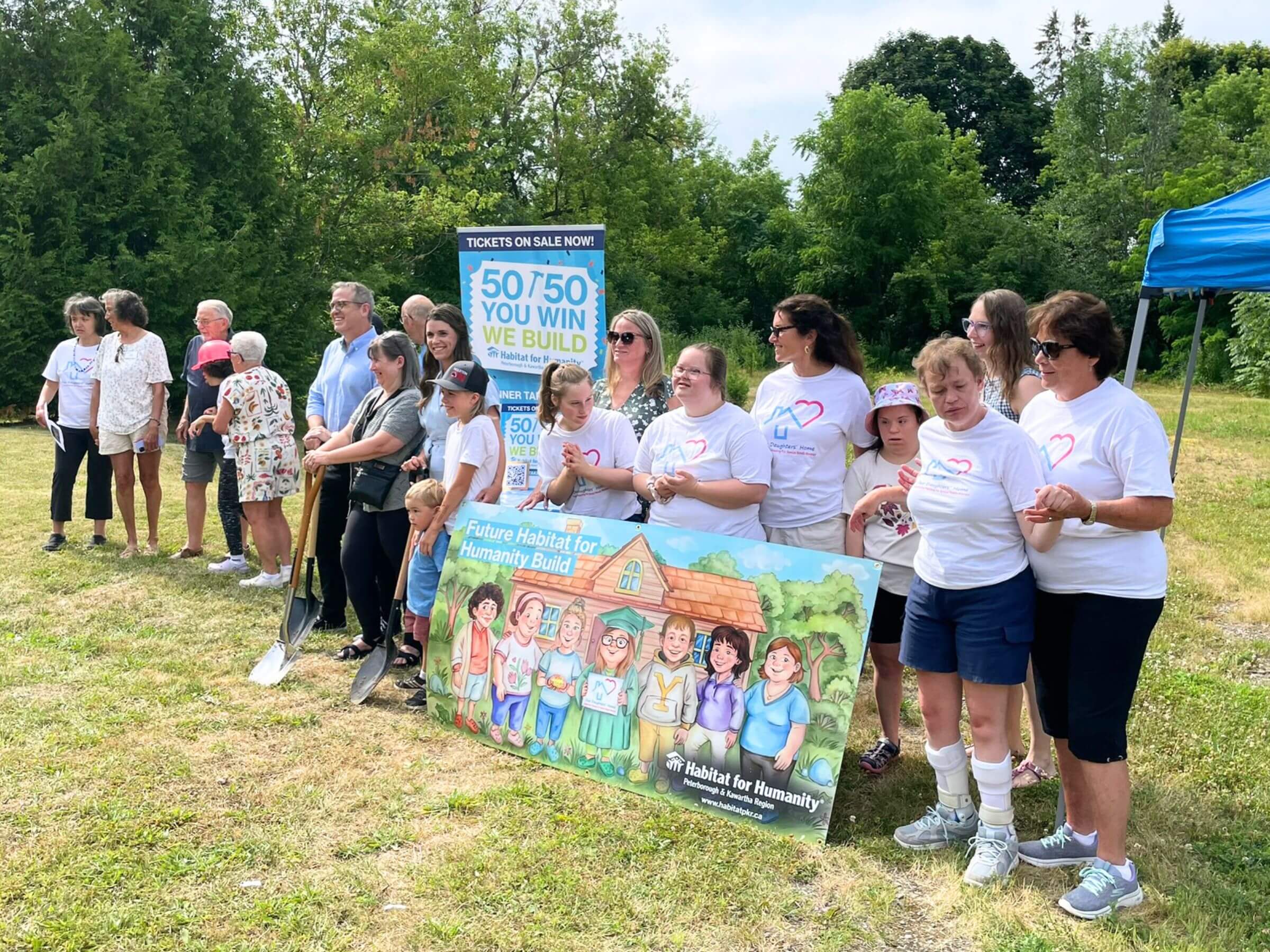 A diverse group of happy people stand outside in a field holding a Habitat branded sign that reads "Future Habitat for Humanity Build". A number of the people are wearing the same "Our Daughter's Home" shirts