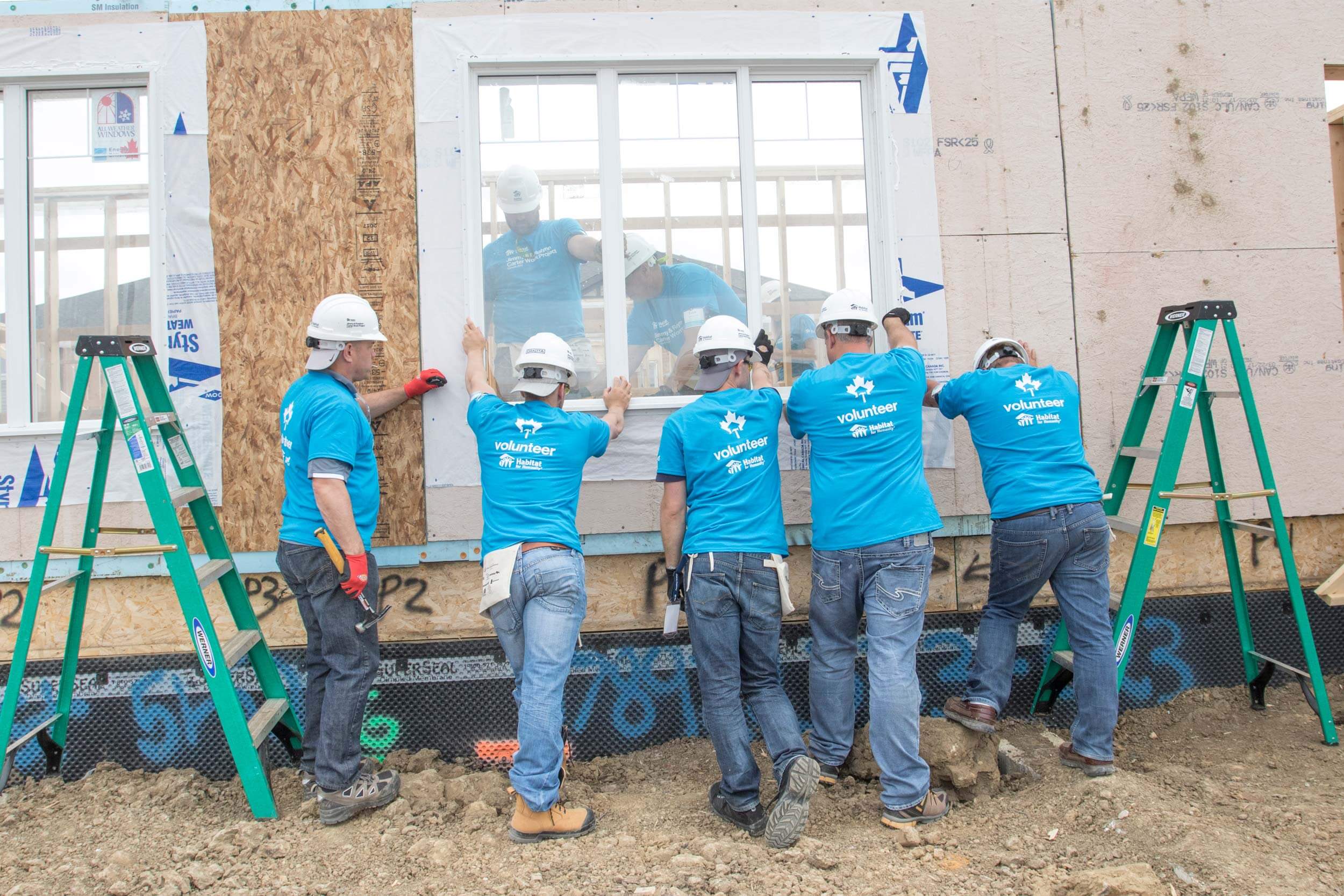 Image of several people wearing blue Habitat for Humanity t-shirts and hard hats holding a window in place on a home that is under construction.