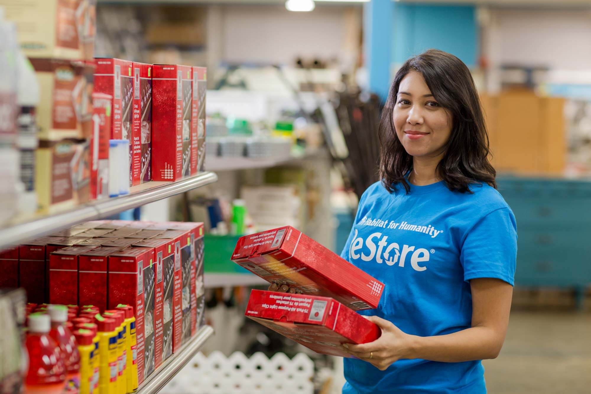 A volunteer wearing a blue ReStore shirt smiles mid task as she's restocking shelves