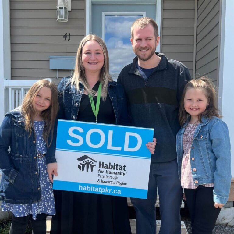 A smiling family of four with 2 young daughters stand in front of their new home, smiling wide holding a Habitat branded "SOLD" sign