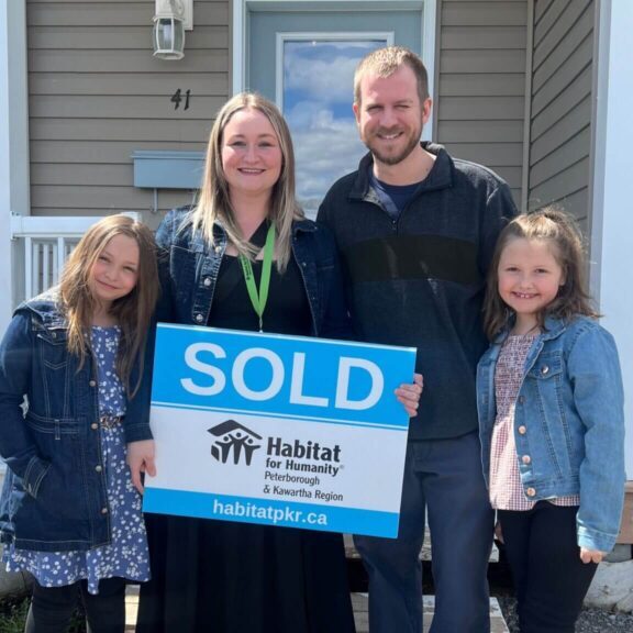 A smiling family of four with 2 young daughters stand in front of their new home, smiling wide holding a Habitat branded "SOLD" sign