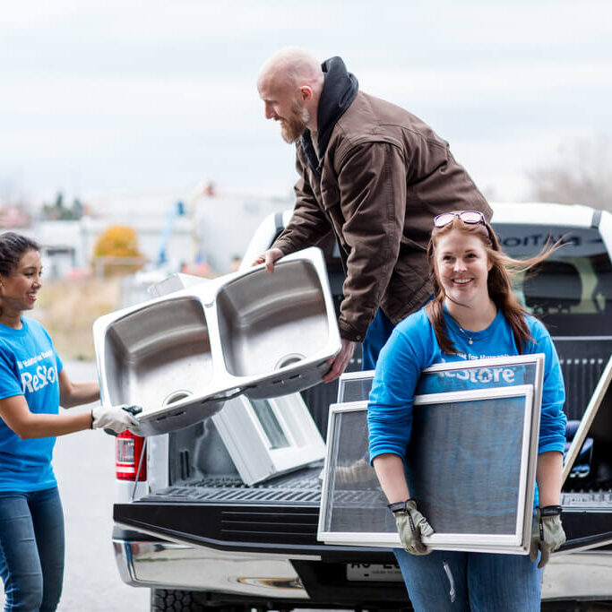 A pickup truck has its tailgate down, a man is standing in the box unloading sinks and windows to two helpful women in blue Habitat ReStore shirts
