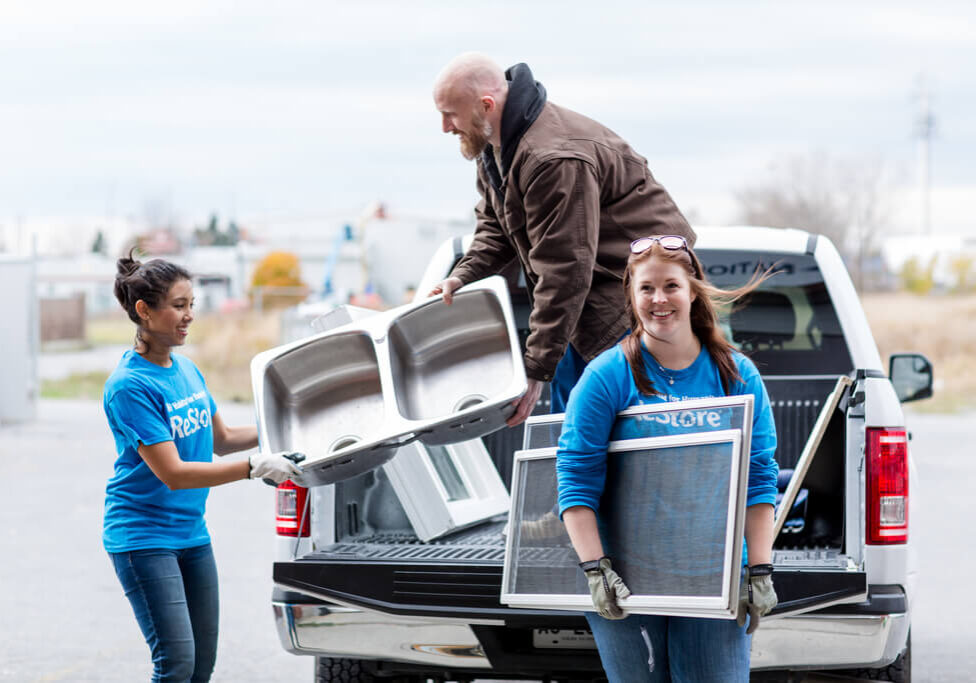 A pickup truck has its tailgate down, a man is standing in the box unloading sinks and windows to two helpful women in blue Habitat ReStore shirts