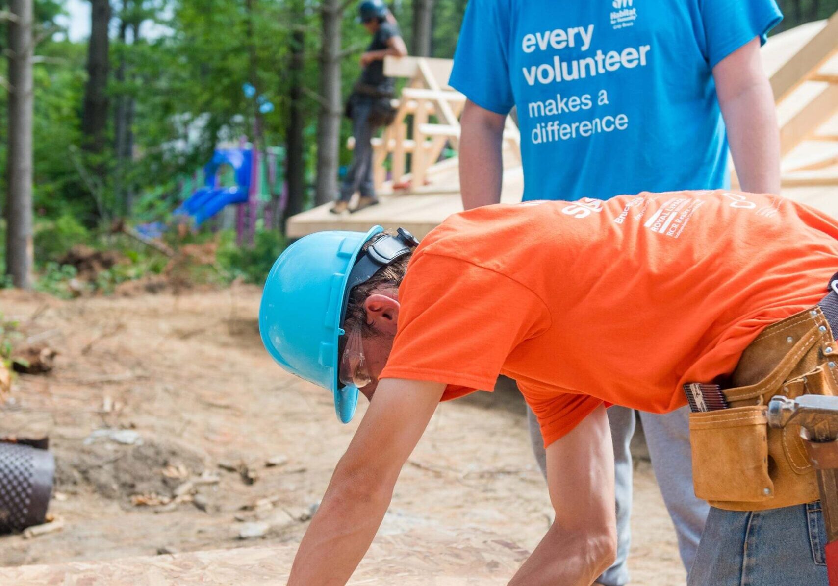 A man in a volunteer shirt and hardhat is making a pencil mark on a piece of plywood alongside his tape measure