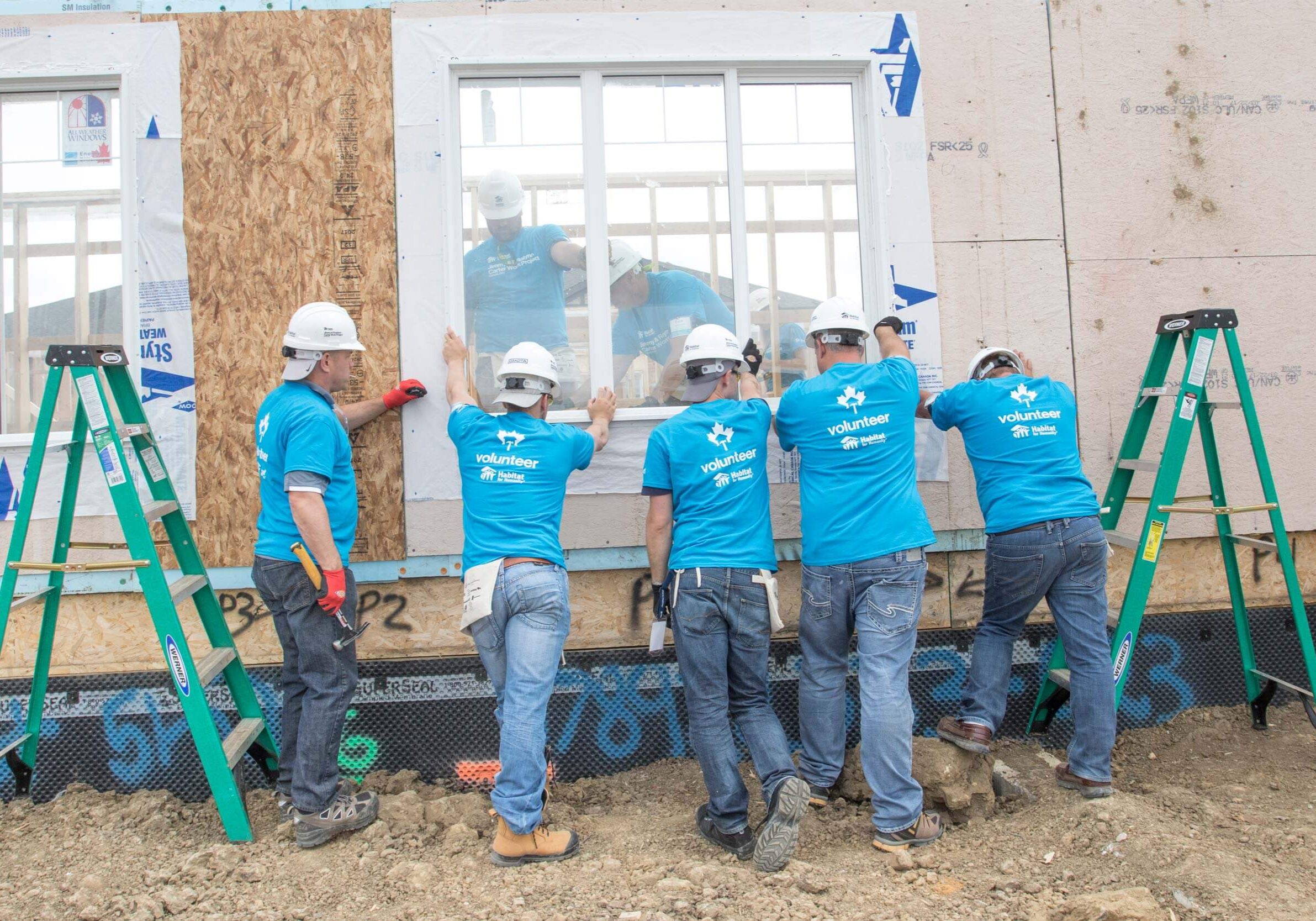 Image of several people wearing blue Habitat for Humanity t-shirts and hard hats holding a window in place on a home that is under construction.