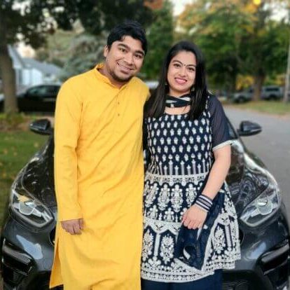 A couple are smiling in front of their neighbourhood street, he is dressed in cheery yellow and she has a beautiful patterned dress