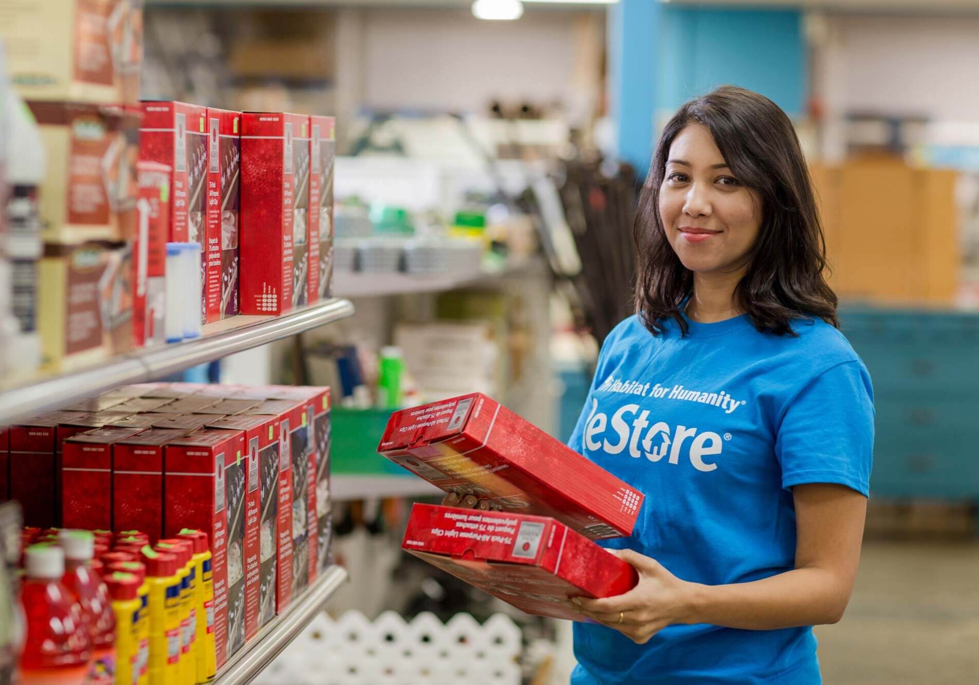 A volunteer wearing a blue ReStore shirt smiles mid task as she's restocking shelves