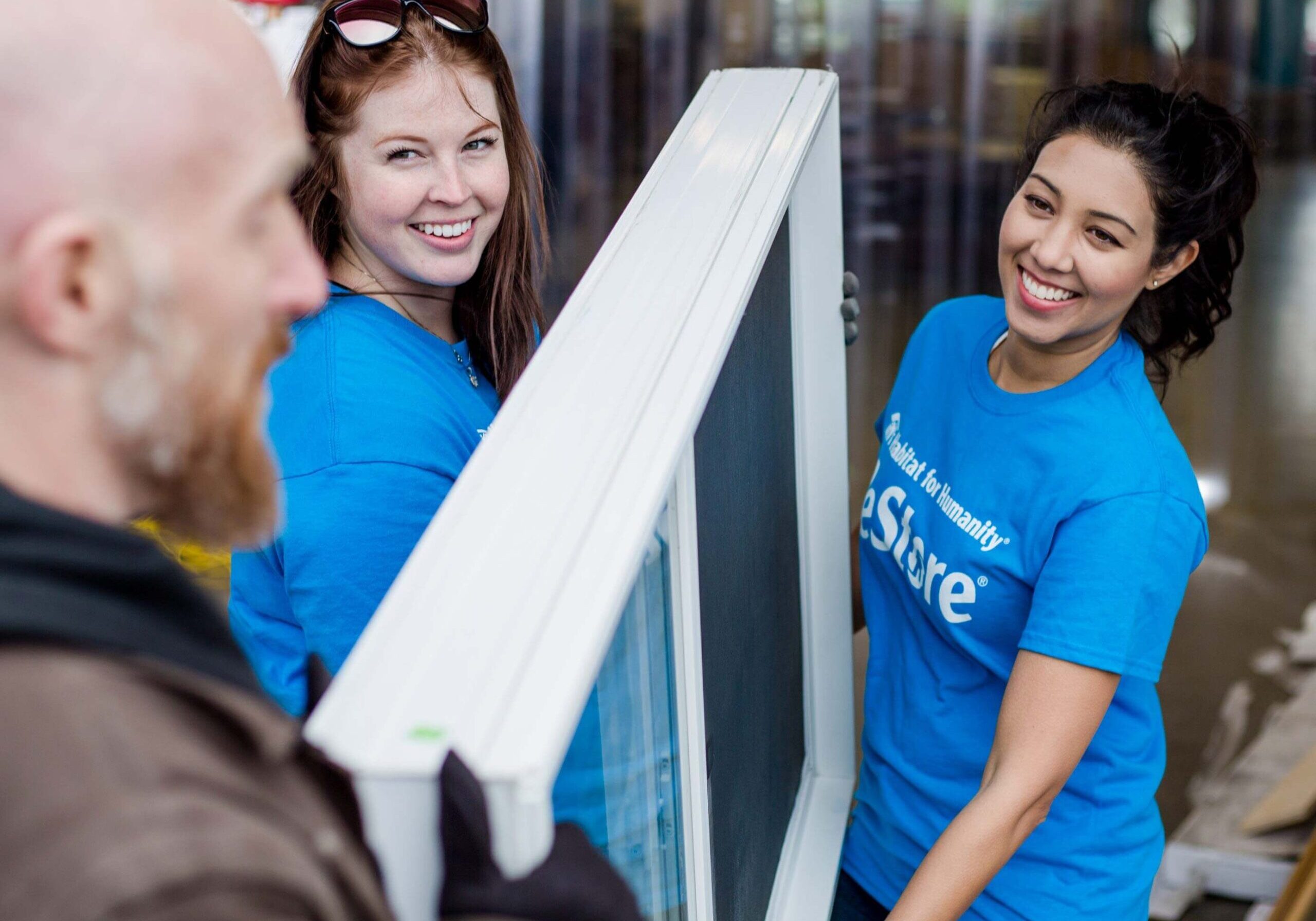 2 smiling volunteers in blue ReStore shirt are happily helping a man carry an oversized donated window into the store