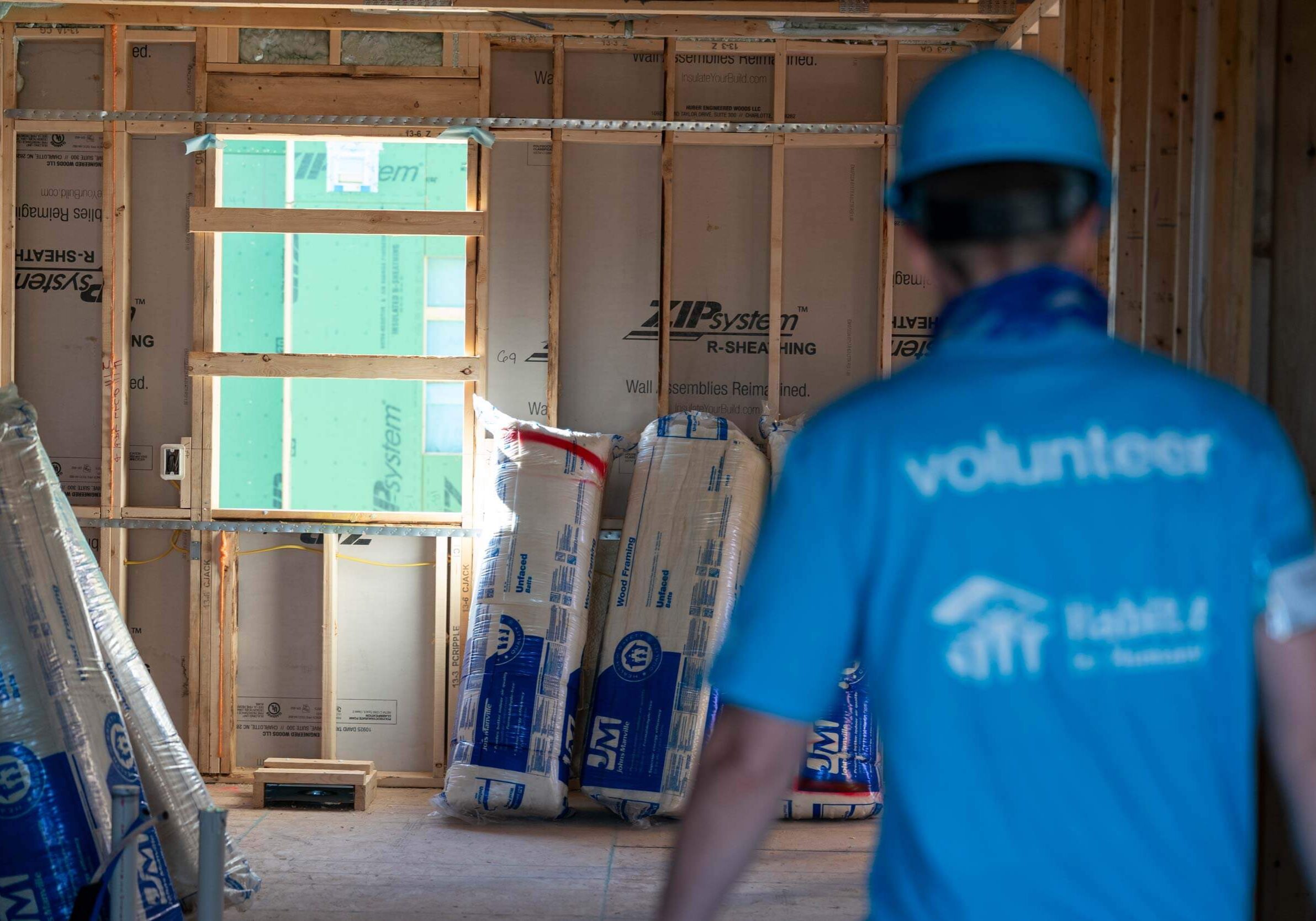 A volunteer in a blue Habitat shirt and hardhat in walking through an under construction home build towards bags of insulation
