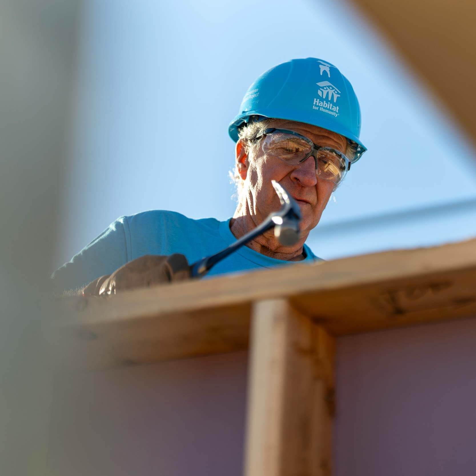 An older man wearing a blue Habitat hardhat is seen through two beams, hammering in a nail