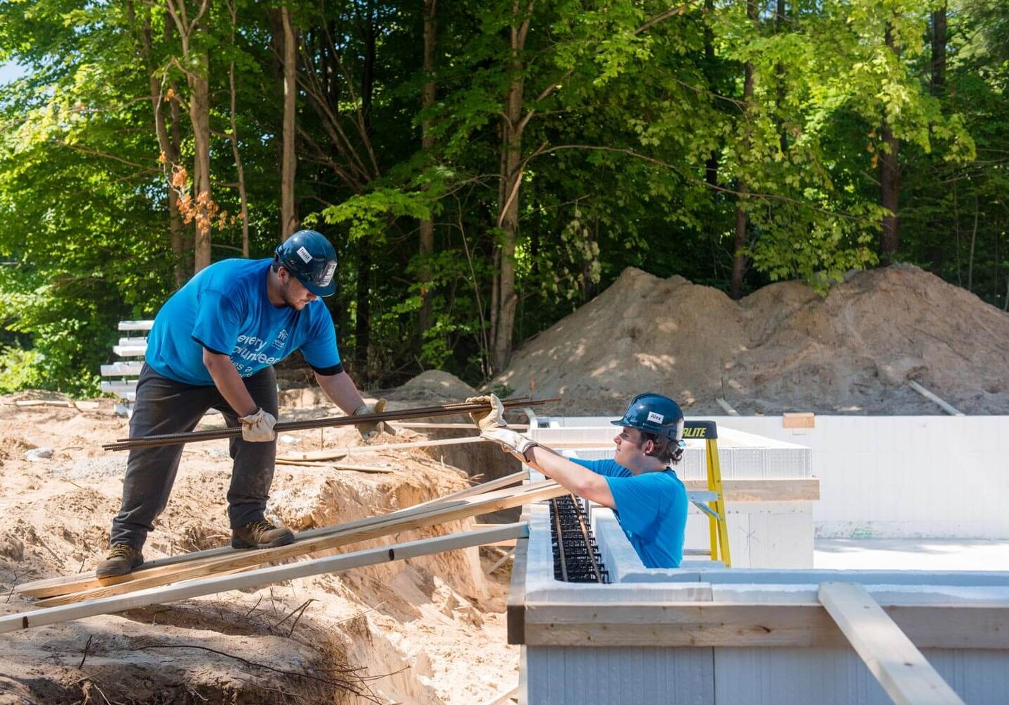 2 men working outside on a build site are handing each other a long piece of wood. They are both in safety gear and Habitat blue volunteer shirts
