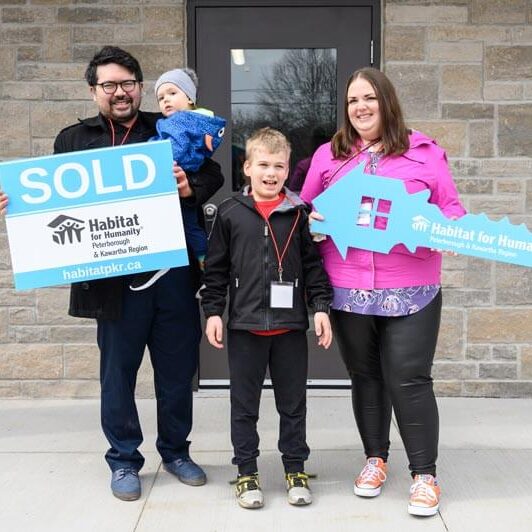 A family of 4, 2 adults and 2 young kids, stand beaming in front of their new home, holding a large Habitat branded SOLD sign and house oversized key cutout