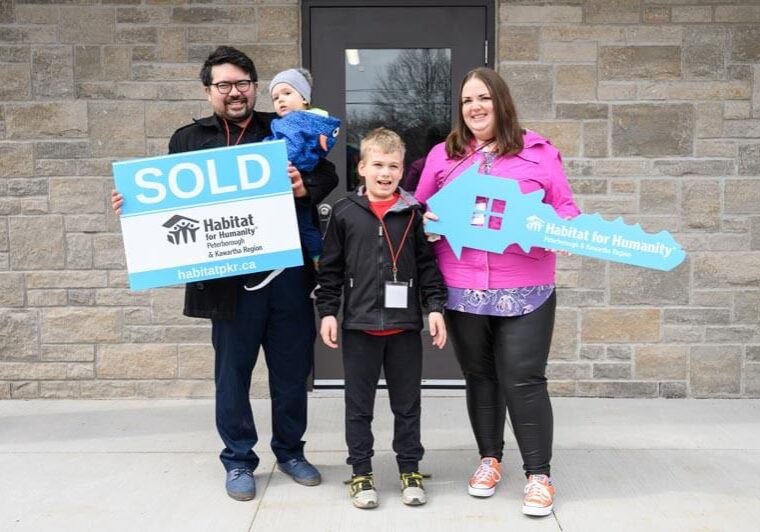 A family of 4, 2 adults and 2 young kids, stand beaming in front of their new home, holding a large Habitat branded SOLD sign and house oversized key cutout